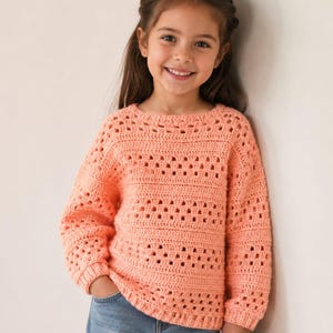 Pode incluir: Uma camisola de croché de manga comprida cor de pêssego com um padrão repetitivo de faixas horizontais sólidas alternadas com filas de pontos quadrados abertos, combinada com calças de ganga azuis. A camisola tem um decote redondo e punhos canelados.