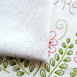 May include: Close-up of white fabric with floral embroidery and a dragonfly outline. The embroidery features pink flowers, green leaves, and stems. The dragonfly and flower outlines are in gray. The text "Remember" is embroidered in brown.