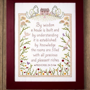 Może przedstawiać: Biały materiał haftowany z czerwoną ramą, przedstawiający dom z zielonym dachem i cytat z Przysłów 24:3-4: "Mądrością buduje się dom, a rozumem umacnia się go: przez poznanie pokoje napełniają się wszelkimi cennymi i przyjemnymi bogactwami."