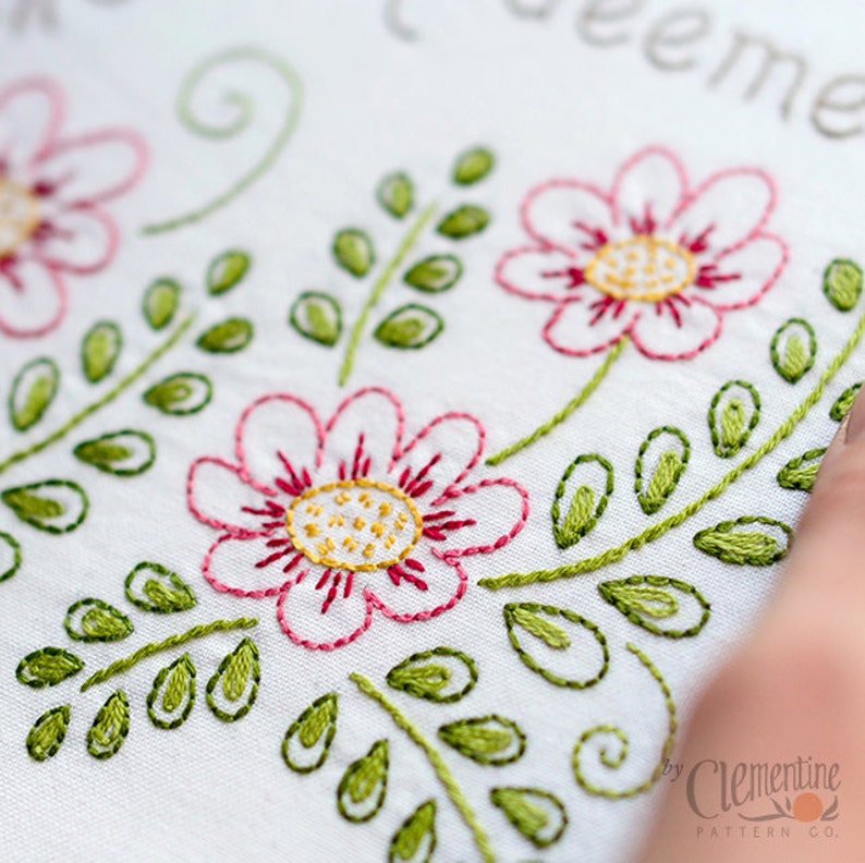 May include: Close-up of white fabric with embroidered floral design. Pink flowers with red outlines and yellow centers are featured, along with green leaves and stems. The embroidery is detailed and delicate, showcasing a handcrafted aesthetic.