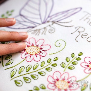 May include: White fabric with embroidered floral and butterfly design. The embroidery features pink flowers with yellow centers, green leaves, and a purple butterfly. The text "The Read" is also embroidered. The design is by Clementine Pattern Co.