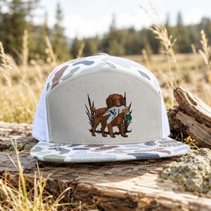 May include: A baseball cap featuring a light gray front panel with an embroidered brown dog holding a mallard duck in its mouth, surrounded by tall grass. The cap has a white mesh back and a brim with a camouflage pattern in shades of gray and green.