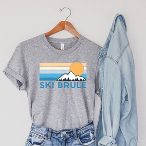 Könnte beinhalten: Graues T-Shirt mit Berg- und Sonnengrafik und dem Text "SKI BRULE" in Blau. Das Shirt wird mit Jeansshorts und einer hellblauen Jeansjacke präsentiert.