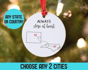 Puede incluir: Adorno navideño blanco y redondo con cinta roja. Muestra un mapa con dos estados unidos por una línea de puntos y corazones. Texto: "ALWAYS close at heart", "ANY STATE OR COUNTRY" y "CHOOSE ANY 2 CITIES".