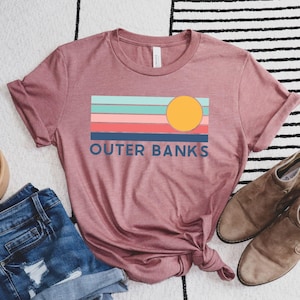 May include: A dusty rose t-shirt with a colorful graphic that reads "Outer Banks." The graphic features a yellow sun and horizontal stripes in shades of blue, pink, and coral. The shirt is paired with denim shorts and brown suede ankle boots.
