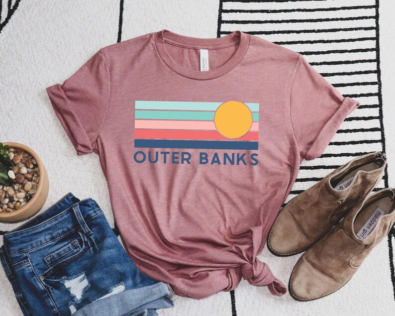 May include: A dusty rose t-shirt with a colorful graphic that reads "Outer Banks." The graphic features a yellow sun and horizontal stripes in shades of blue, pink, and coral. The shirt is paired with denim shorts and brown suede ankle boots.