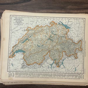 Puede incluir: Una pila de mapas antiguos con una ilustración detallada de Suiza. El mapa está en tonos beige, verde y marrón, con acentos azules para lagos y ríos. El título "RAND MCNALLY SWITZERLAND" es visible.