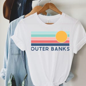 May include: White t-shirt with a colorful graphic design featuring horizontal stripes in teal, light pink, coral, and navy blue, with a yellow circle representing the sun. The text "OUTER BANKS" is printed in navy blue below the stripes.