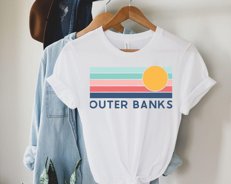 May include: White t-shirt with a colorful graphic design featuring horizontal stripes in teal, light pink, coral, and navy blue, with a yellow circle representing the sun. The text "OUTER BANKS" is printed in navy blue below the stripes.