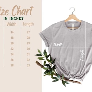 May include: A light gray t-shirt displayed on a wooden hanger, with a size chart in inches. The chart lists sizes XS to 3XL, with corresponding width and length measurements. Greenery accents the shirt.