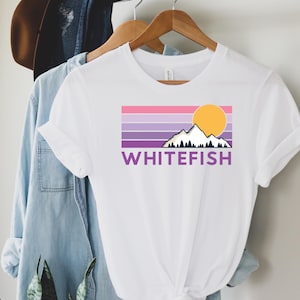 May include: White t-shirt featuring a mountain and sun graphic with the word "WHITEFISH" in purple. The graphic has a retro design with horizontal stripes in shades of pink, purple, and lavender. The shirt is hanging on a wooden hanger.