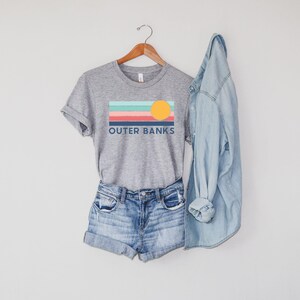 May include: A heather gray t-shirt with the text "OUTER BANKS" and a colorful graphic of a sunset. The shirt is paired with denim shorts and a light blue denim jacket, all hanging on a wooden hanger.