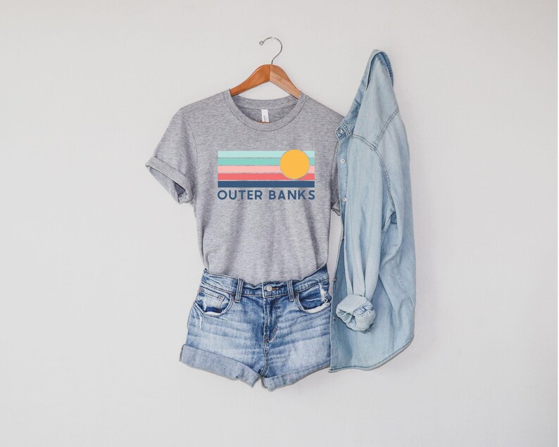 May include: A heather gray t-shirt with the text "OUTER BANKS" and a colorful graphic of a sunset. The shirt is paired with denim shorts and a light blue denim jacket, all hanging on a wooden hanger.