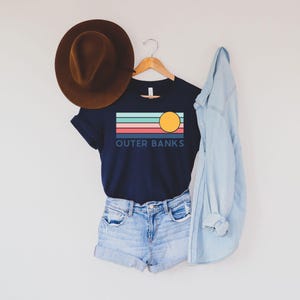 May include: A navy blue t-shirt with the text "OUTER BANKS" and a graphic of a sun and stripes in teal, coral, and light blue. The shirt is paired with denim shorts, a brown hat, and a light blue denim jacket.