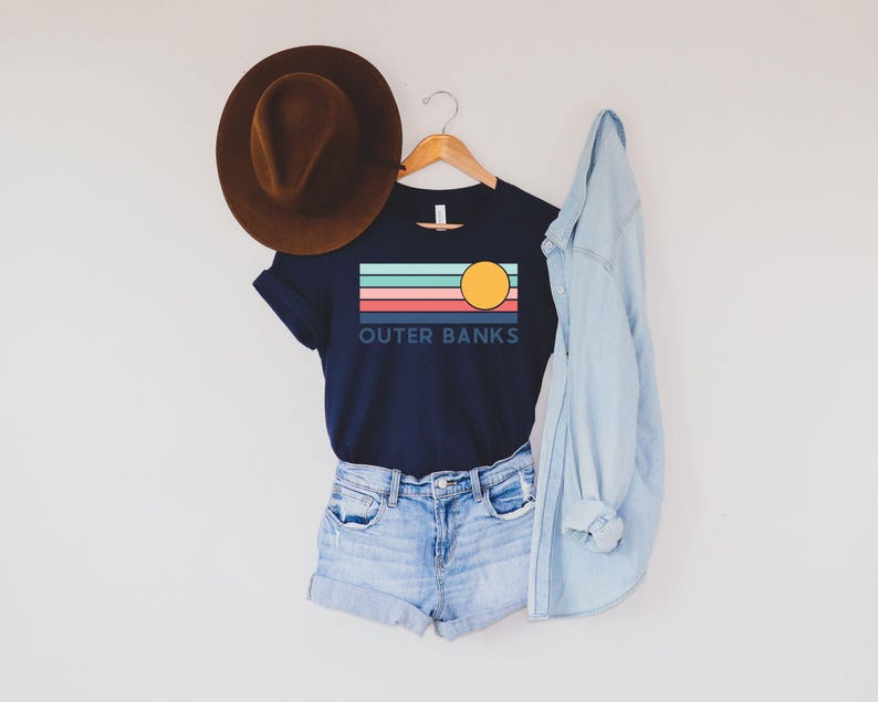 May include: A navy blue t-shirt with the text "OUTER BANKS" and a graphic of a sun and stripes in teal, coral, and light blue. The shirt is paired with denim shorts, a brown hat, and a light blue denim jacket.