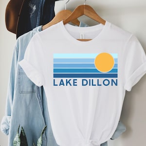 Pode incluir: Camiseta branca com um design gráfico com listras azuis horizontais, um sol amarelo e o texto "LAKE DILLON" em azul. A camiseta está pendurada em um cabide de madeira com uma camisa jeans e um chapéu no fundo.