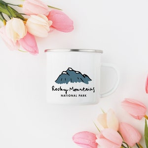 May include: White enamel mug with a black and blue illustration of mountains and the text "Rocky Mountains National Park".