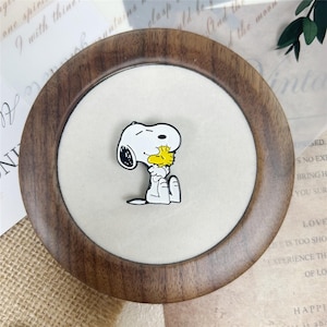 Peut inclure: Une broche en émail blanc et noir représentant le personnage de dessin animé Snoopy serrant dans ses bras un petit oiseau jaune. La broche a une finition lisse avec des contours noirs définis et est centrée sur une surface de présentation circulaire.