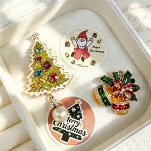 Può includere: Una spilla a forma di albero di Natale dorato con cristalli colorati e una perla bianca pendente, accanto a una spilla con agrifoglio e decorazioni in smalto rosso, verde e oro con un cristallo centrale, vicino a due adesivi circolari con testo.