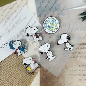 Puede incluir: Una colección de seis pines de esmalte que muestran al personaje de dibujos animados Snoopy en varias poses, incluyendo leyendo un libro, sentado en un banco y de pie. Un pin circular presenta el texto EMOTIONAL SUPPORT ANIMAL con un perro.