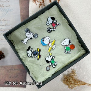 May include: A set of Snoopy character pins in a dark green box. Each pin features Snoopy in different sports and activities, including weightlifting, soccer, surfing, basketball, skateboarding, and tennis. The text "Gift for Anime Fans" is at the bottom.