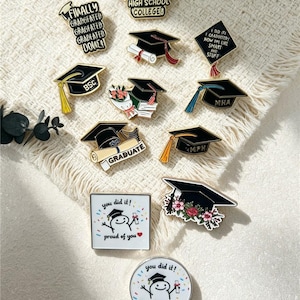 Könnte beinhalten: Eine Sammlung von Emaille-Anstecknadeln zum Thema Abschlussfeier, darunter schwarze Doktorhüte mit Quasten, Diplome, Blumenmotive sowie weiße Anstecker mit Comicfiguren und dem Text you did it! proud of you und verschiedenen Abkürzungen.