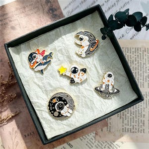 May include: A set of five enamel pins featuring space-themed designs including an astronaut riding a whale, an astronaut on a crescent moon, an astronaut holding a yellow star, an astronaut sitting on a planet with rings, and a circular moon scene.