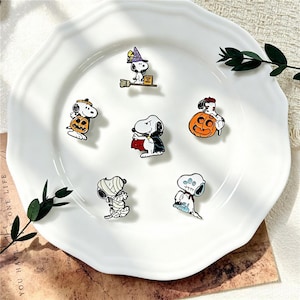 Peut inclure: Un ensemble de six broches en émail représentant le personnage de dessin animé Snoopy dans divers costumes d'Halloween, incluant une sorcière, un vampire, une momie, un fantôme et des personnages interagissant avec des citrouilles orange.