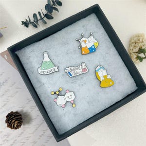 Peut inclure: Un ensemble de cinq broches en émail avec des motifs sur le thème de la science, incluant une fiole verte marquée SCIENCE, une fiole jaune avec un visage, une structure moléculaire, des béchers et une bannière avec le texte Science Rules.