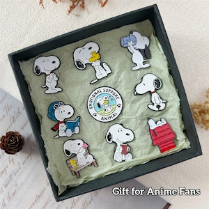 Peut inclure: Une collection d'épingles de personnages Snoopy dans une boîte cadeau noire. Les épingles présentent diverses poses de Snoopy et Woodstock, ainsi qu'une épingle "Emotional Support Animal". Le texte "Gift for Anime Fans" est visible.