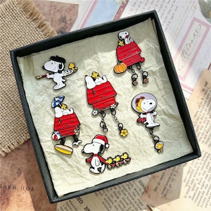 Può includere: Una collezione di sei spille in smalto raffiguranti Snoopy e Woodstock in vari temi stagionali, tra cui un pittore, un pilota e personaggi con cucce rosse, zucche e accessori festivi, disposti all'interno di una scatola quadrata nera.