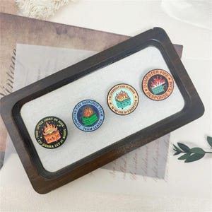 May include: Four circular enamel pins featuring dumpster fire illustrations. The pins display text: "THIS LITTLE LIGHT OF MINE I'M GONNA LET IT SHINE", "DUMPSTER FIRE RESPONSE TEAM LEADER", "THE STRONGEST STEEL IS FORGED IN THE FIRE OF A DUMPSTER", and "DUMPSTER FIRE RESPONSE TEAM".