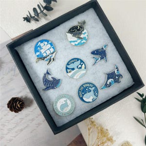Könnte beinhalten: Eine Sammlung von Emaille-Anstecknadeln mit Meeresmotiven. Designs umfassen Wale, Haie und einen Mantarochen, mit Details von Meereswellen und Sternennächten. Die Anstecknadeln werden in einer schwarzen Box mit weißem Innenraum präsentiert.