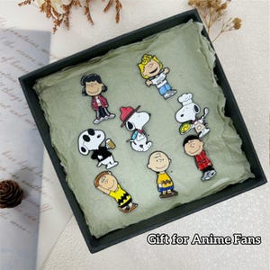 Può includere: Una collezione di spille colorate di personaggi dei cartoni animati, tra cui Snoopy e Charlie Brown, presentate in una scatola regalo nera con interno verde chiaro. Il testo "Gift for Anime Fans" è visibile.