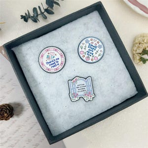 Peut inclure: Trois broches en émail aux motifs pastel. Une broche circulaire porte l'inscription 'there's no need to rush' avec une fraise. Une autre broche circulaire indique 'Small steps are okay too' avec des fleurs. Une broche en forme de fenêtre dit 'don't lose this moment searching for another'.