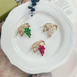 Peut inclure: Trois broches en métal doré en forme de grappes de raisin. Chaque broche présente des feuilles dorées texturées et une grappe de perles rondes. Une broche a des perles vertes, une des perles blanches nacrées et une des perles rouge foncé.