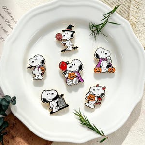 May include: A set of six enamel pins featuring the cartoon character Snoopy in various Halloween costumes, including a witch, a vampire, and a devil, holding items like pumpkins, a red balloon, a lollipop, and standing by a tombstone.