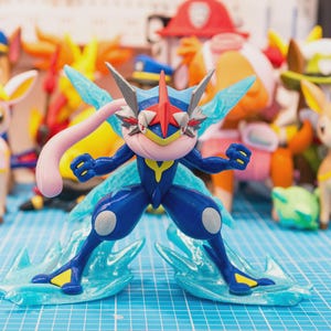 May include: A Greninja action figure featuring a dark blue body with light blue accents, a pink tongue scarf, and a red star-shaped crest on its head. The figure stands on translucent blue water-like base effects with sharp, pointed edges.