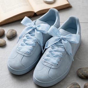 Op de afbeelding: Een paar lichtblauwe suède sneakers met brede, lichtblauw-wit geblokte lintveters die bovenaan tot grote strikken zijn gestrikt. De sneakers hebben een ronde neus, bijpassende blauwe zolen en een gladde, matte afwerking.