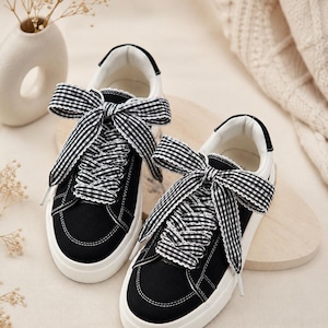 May include: A pair of black canvas sneakers featuring white rubber soles and wide black and white gingham patterned ribbon laces tied into large bows at the top. The sneakers have white interior lining and white stitching details along the edges.