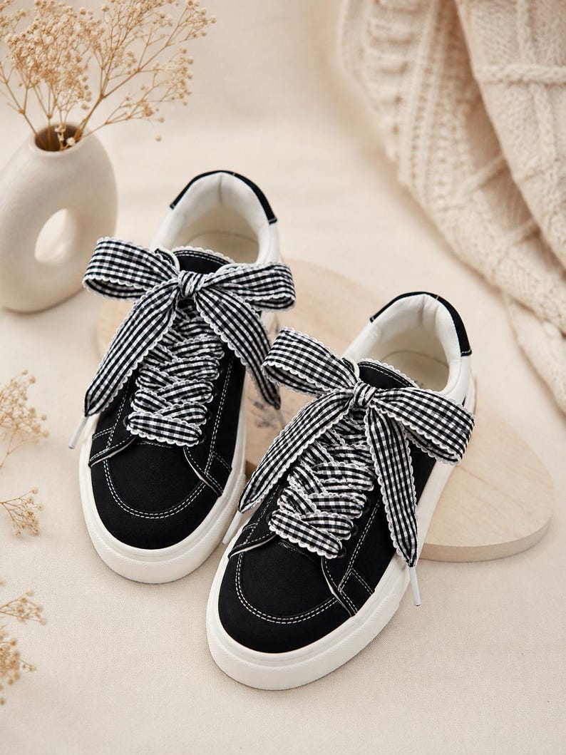 May include: A pair of black canvas sneakers featuring white rubber soles and wide black and white gingham patterned ribbon laces tied into large bows at the top. The sneakers have white interior lining and white stitching details along the edges.