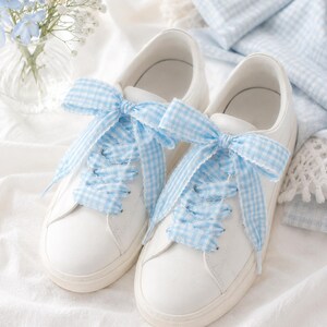 May include: A pair of white low-top sneakers featuring wide, light blue and white gingham patterned ribbon laces tied into large bows at the top. The sneakers have a smooth white exterior, rounded toes, and matching white rubber soles.