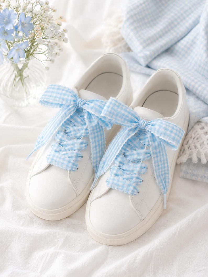 May include: A pair of white low-top sneakers featuring wide, light blue and white gingham patterned ribbon laces tied into large bows at the top. The sneakers have a smooth white exterior, rounded toes, and matching white rubber soles.