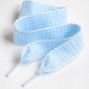 May include: A light blue and white gingham patterned shoelace featuring a scalloped edge trim along both sides and finished with clear plastic aglets at each end. The flat fabric lace is coiled in a loose, layered loop formation.