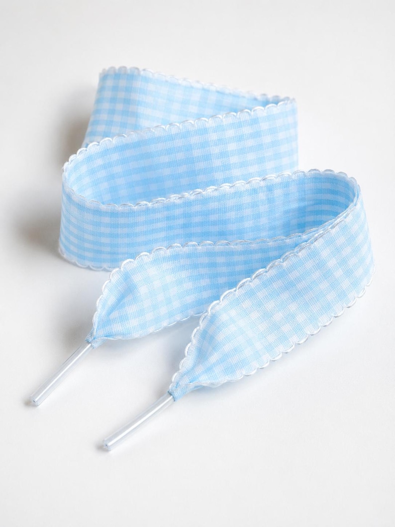 May include: A light blue and white gingham patterned shoelace featuring a scalloped edge trim along both sides and finished with clear plastic aglets at each end. The flat fabric lace is coiled in a loose, layered loop formation.