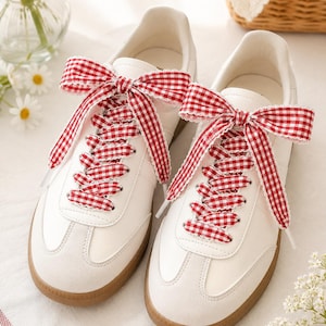 May include: A pair of white sneakers featuring a gum rubber sole and red and white gingham patterned ribbon laces tied into large bows at the top. The shoes have a smooth white upper with suede-like overlays at the toe and lace eyelet panels.