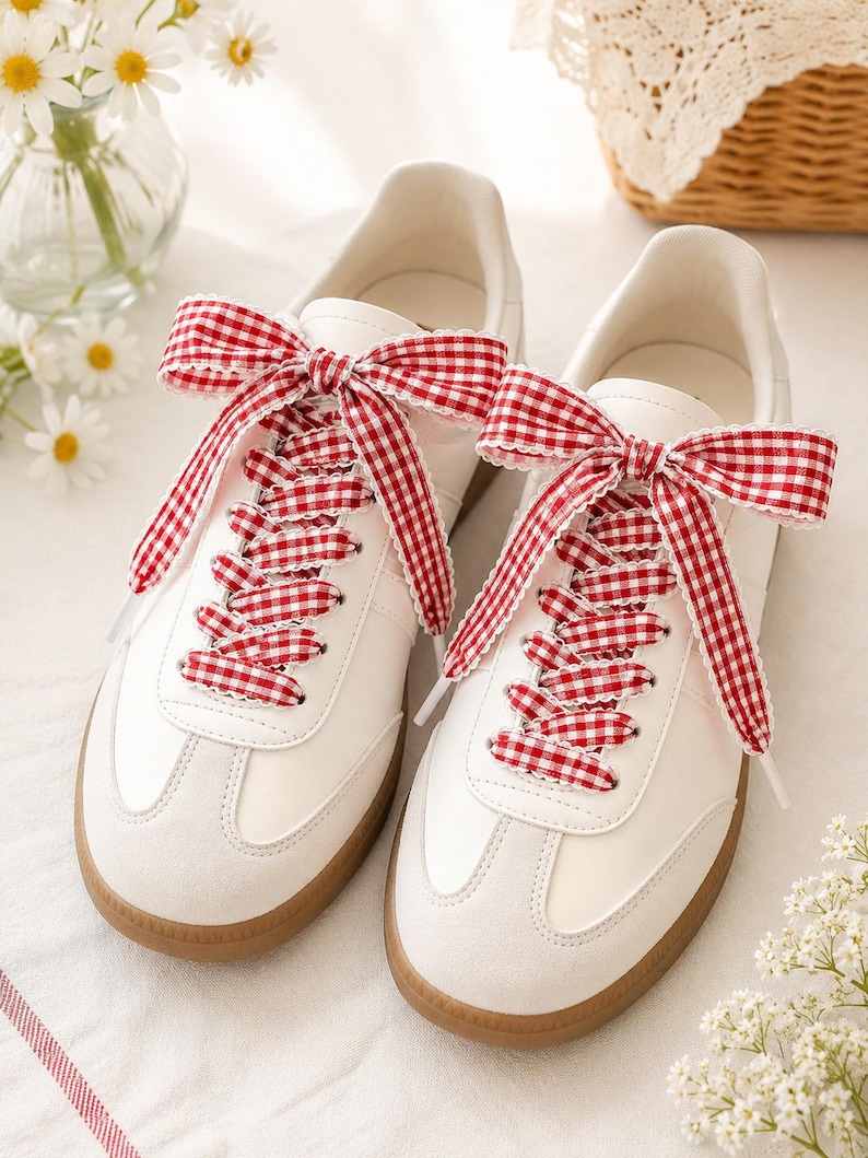 May include: A pair of white sneakers featuring a gum rubber sole and red and white gingham patterned ribbon laces tied into large bows at the top. The shoes have a smooth white upper with suede-like overlays at the toe and lace eyelet panels.
