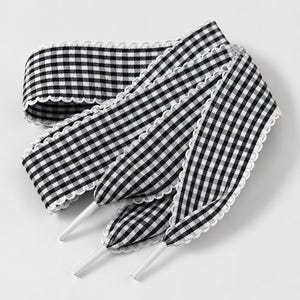 May include: A pair of flat shoelaces featuring a black and white gingham check pattern. The laces have a scalloped white trim along both edges and are finished with solid white plastic aglets at the ends for easy threading through eyelets.