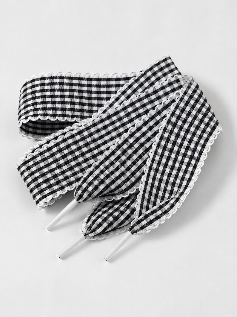 May include: A pair of flat shoelaces featuring a black and white gingham check pattern. The laces have a scalloped white trim along both edges and are finished with solid white plastic aglets at the ends for easy threading through eyelets.