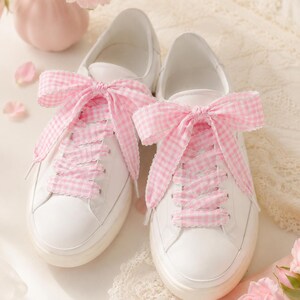 May include: A pair of white low-top sneakers featuring wide, light pink and white gingham patterned fabric laces tied into large bows at the top. The sneakers have a smooth white leather-like exterior, rounded toes, and thick white rubber soles.